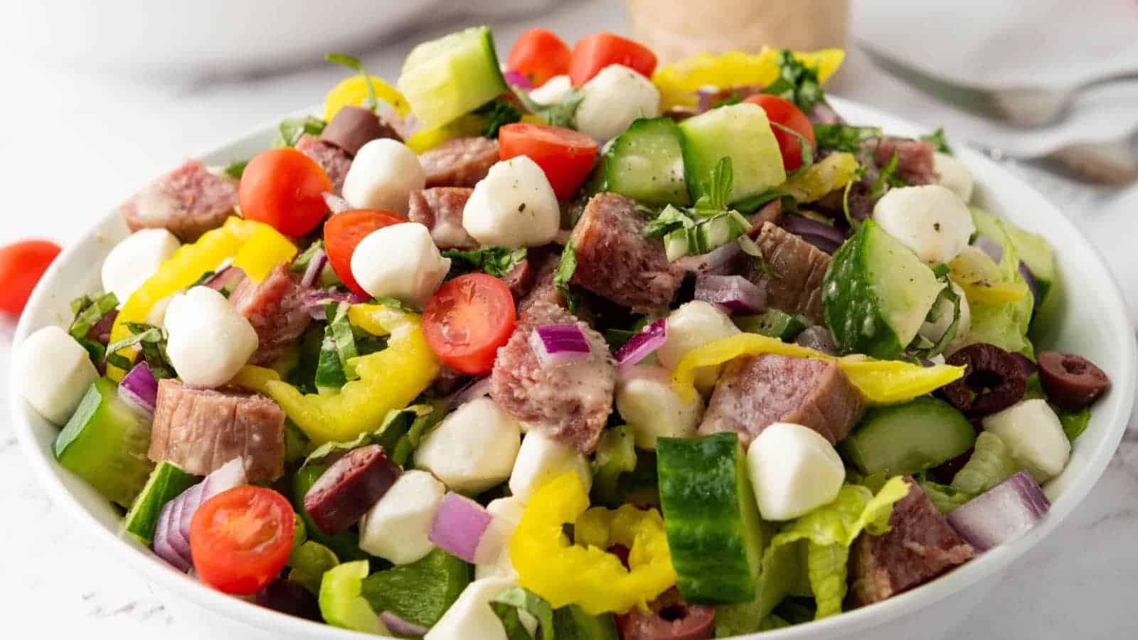 A bowl of chopped salad with steak, cherry tomatoes, cucumber, mozzarella balls, red onion, olives, and yellow peppers.