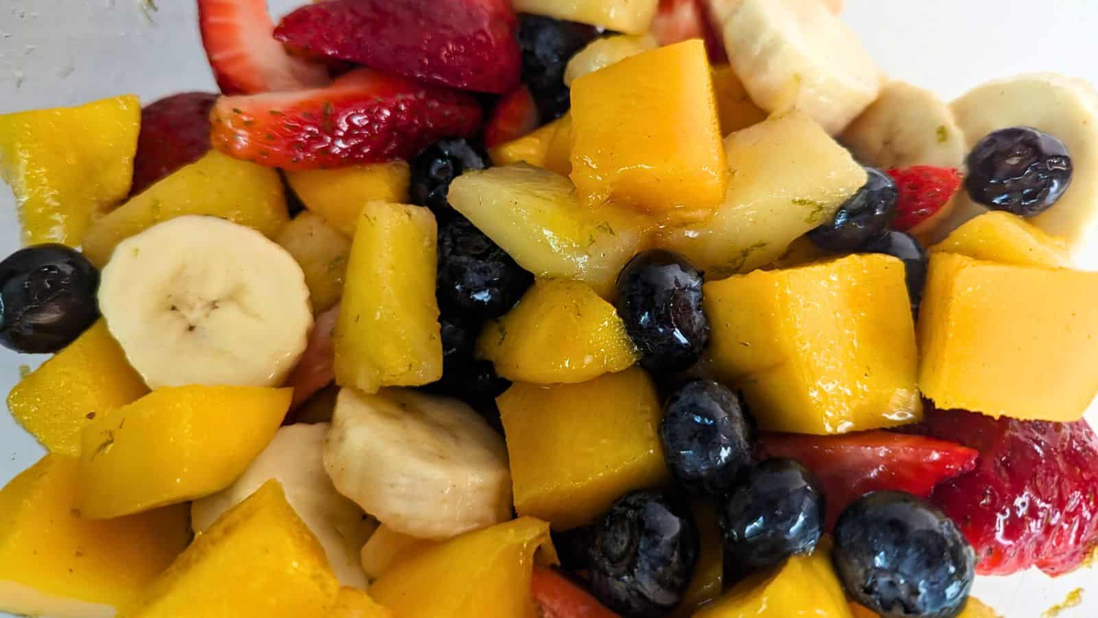 A close-up of a mixed fruit salad containing sliced bananas, strawberries, mangoes, blueberries, and pineapples.