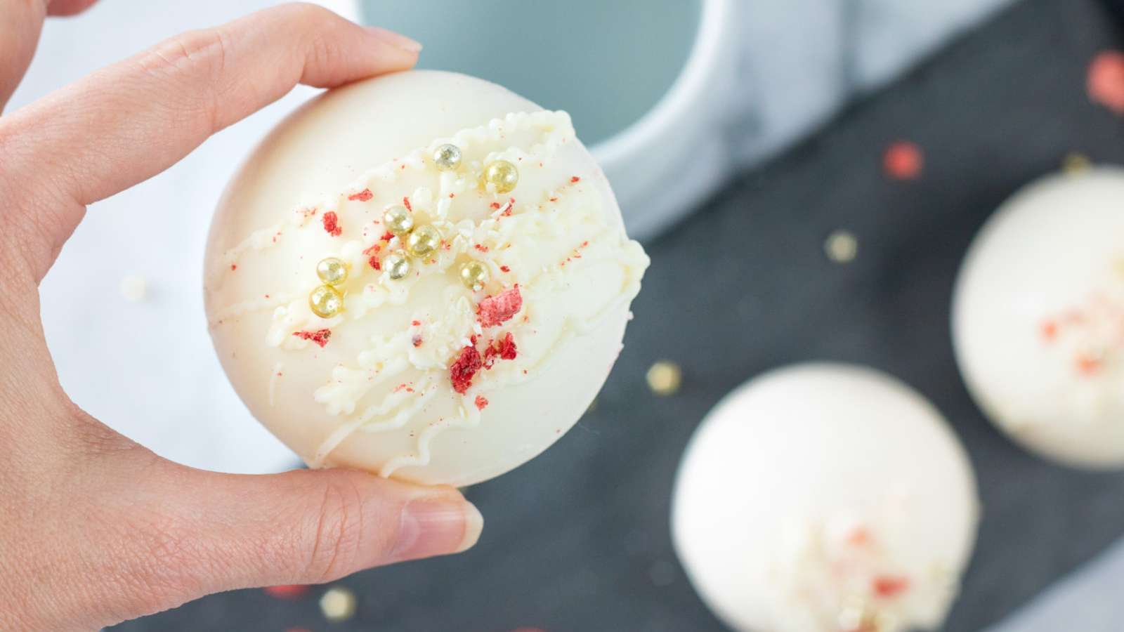 A hand holds a decorated white chocolate sphere topped with white drizzle, gold pearls, and red crumbs; similar spheres are on a black tray below.