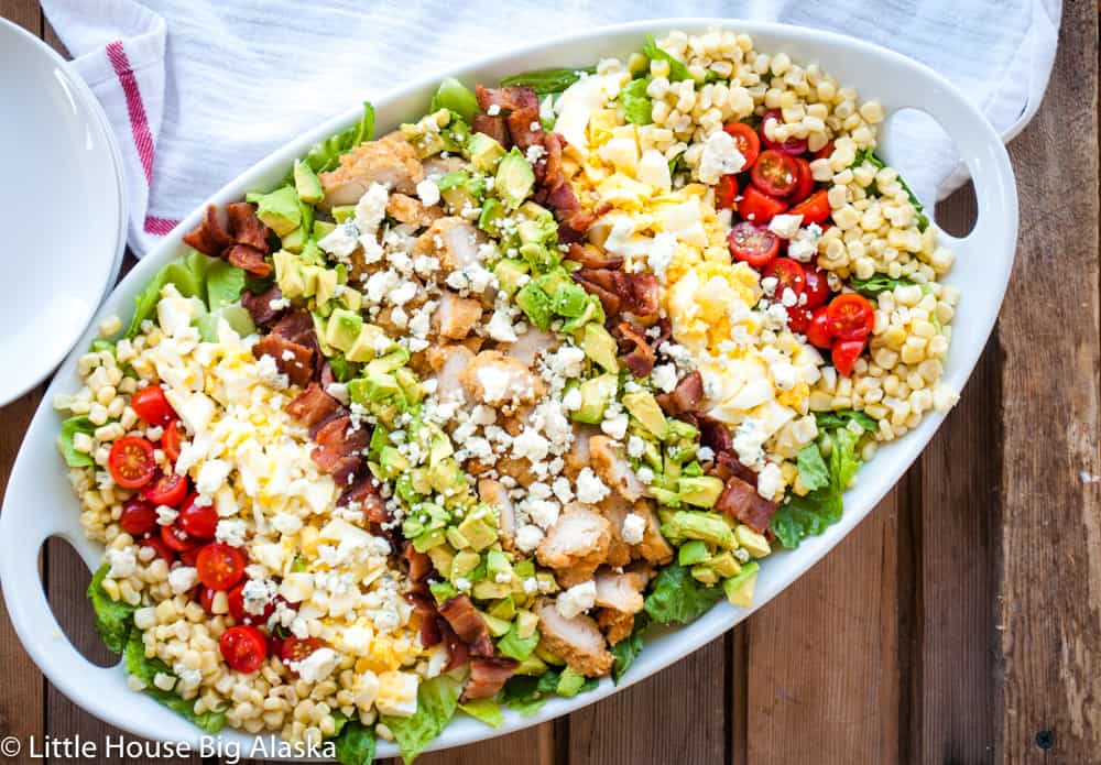 Oval platter with rows of chopped chicken, bacon, avocado, tomatoes, corn, hard-boiled eggs, and crumbled cheese on a bed of lettuce.