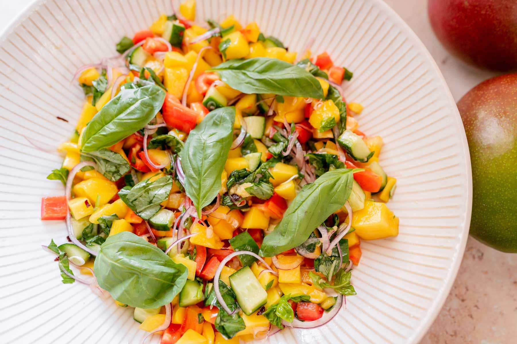 A plate of mango salad with diced mango, cucumber, red bell pepper, basil leaves, and thinly sliced red onion on a white dish.