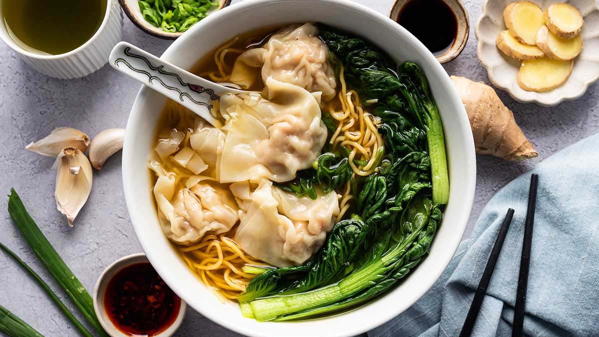 Bowl of wonton noodle soup with dumplings, leafy greens, and broth, surrounded by fresh ingredients and condiments on a table.