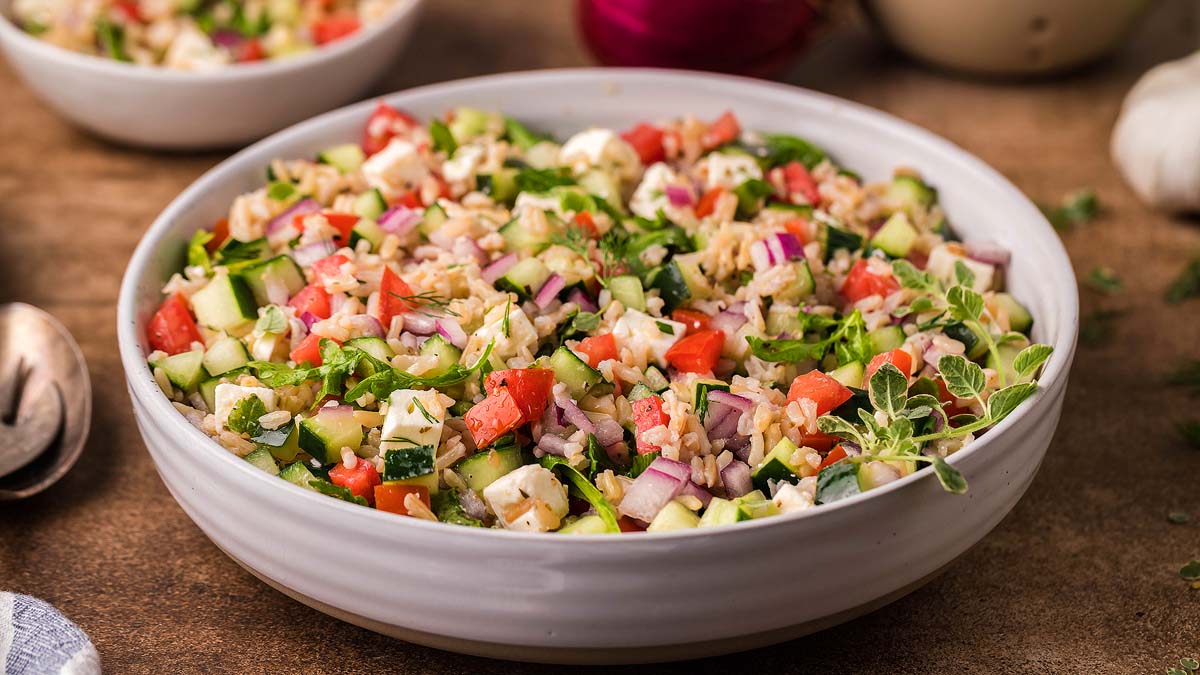 A white bowl filled with a mixed salad containing chopped cucumbers, tomatoes, red onions, greens, and grains, set on a brown surface.