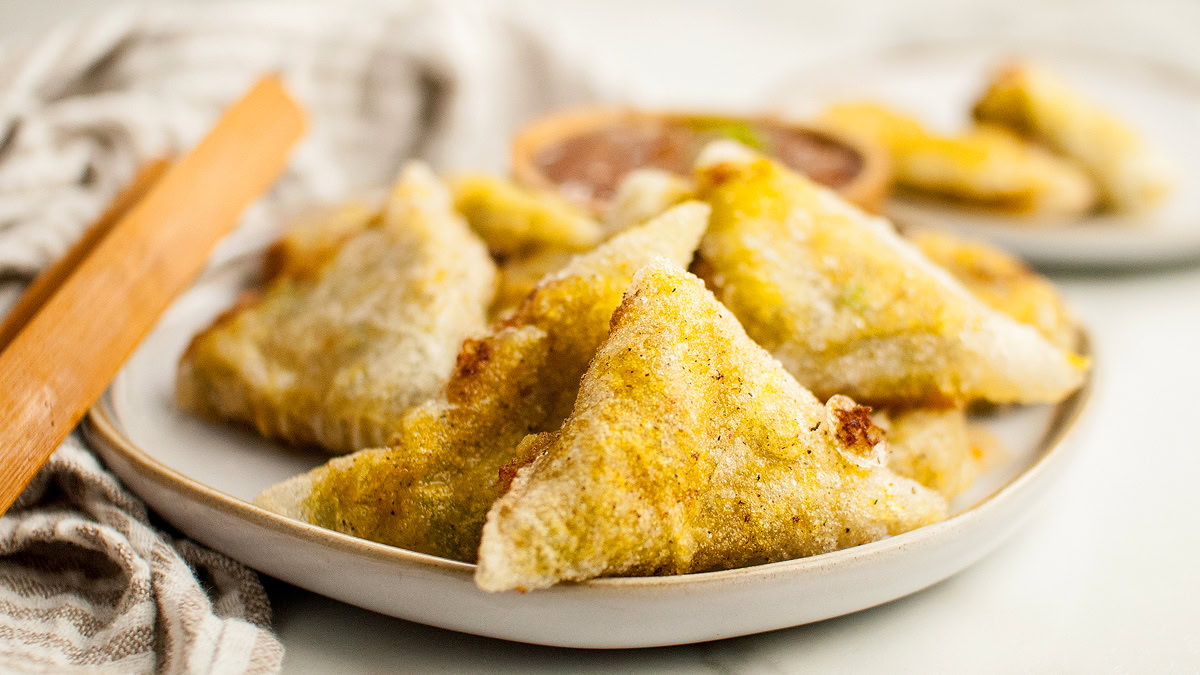 A plate of golden, crispy, triangular samosas is served with a small bowl of dipping sauce in the background.