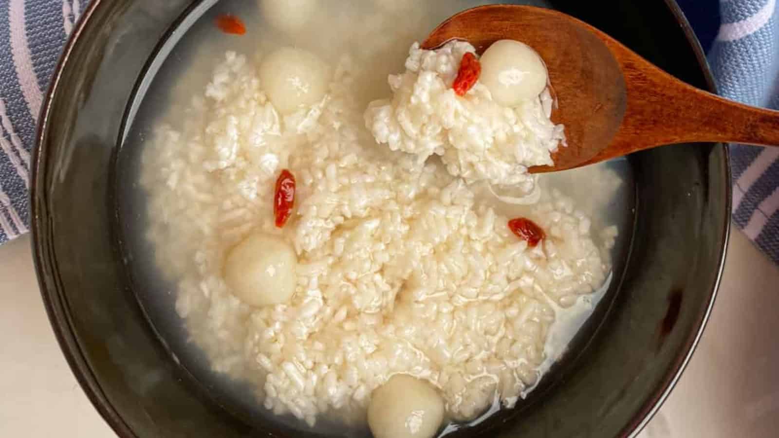 A bowl of sweet fermented rice soup with small white glutinous rice balls and goji berries, with a wooden spoon scooping some from the bowl.