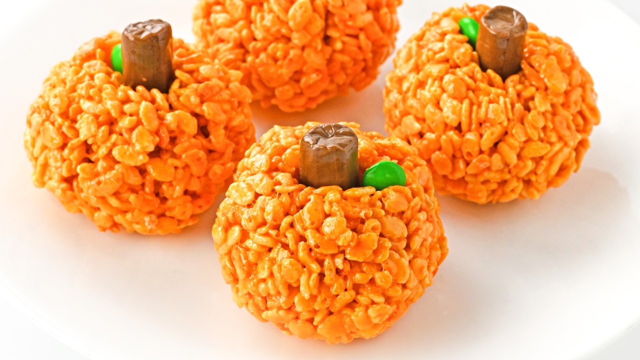 Four pumpkin-shaped Rice Krispies treats with orange coloring, chocolate pieces as stems, and green candies as leaves are arranged on a white plate.