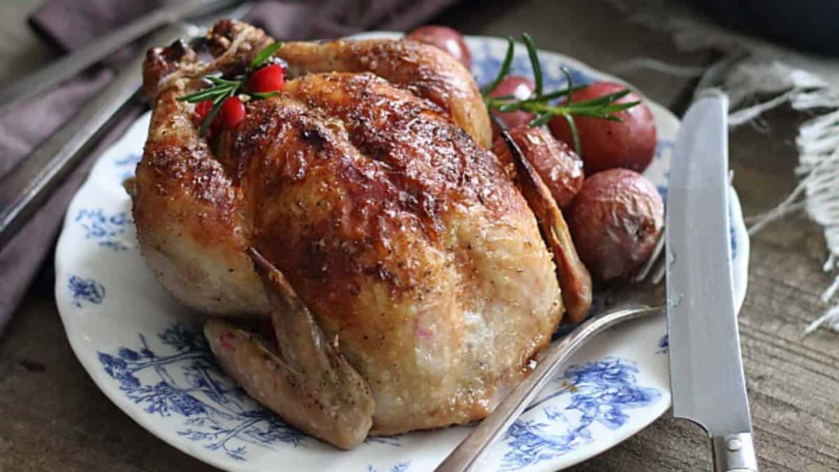 A roasted whole chicken garnished with herbs and berries is served on a blue-and-white plate next to roasted potatoes, with a knife and serving fork beside the plate.