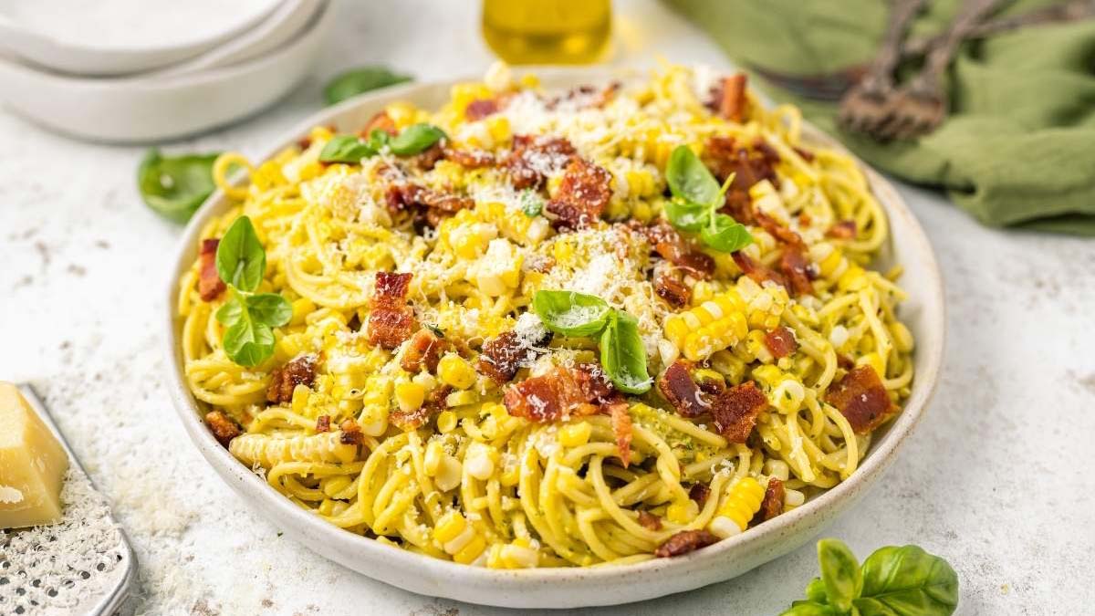 A plate of spaghetti topped with corn, bacon pieces, grated cheese, and fresh basil leaves, set on a light-colored surface with utensils and a green napkin in the background.
