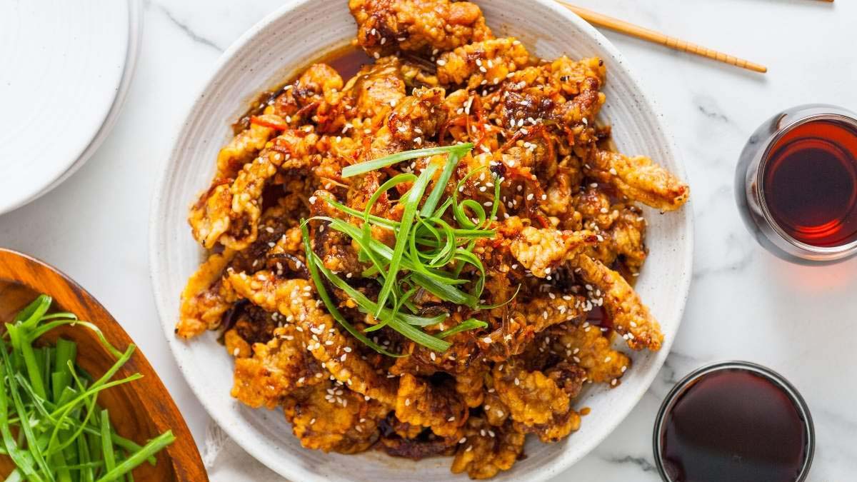 A plate of crispy fried meat strips topped with sesame seeds and sliced green onions, served with two glasses of dark-colored beverage.