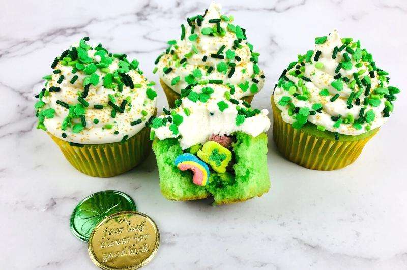 Four cupcakes with green and white sprinkles; one cupcake cut open showing green cake and marshmallow shapes. Two gold coins lie nearby on a white marble surface.