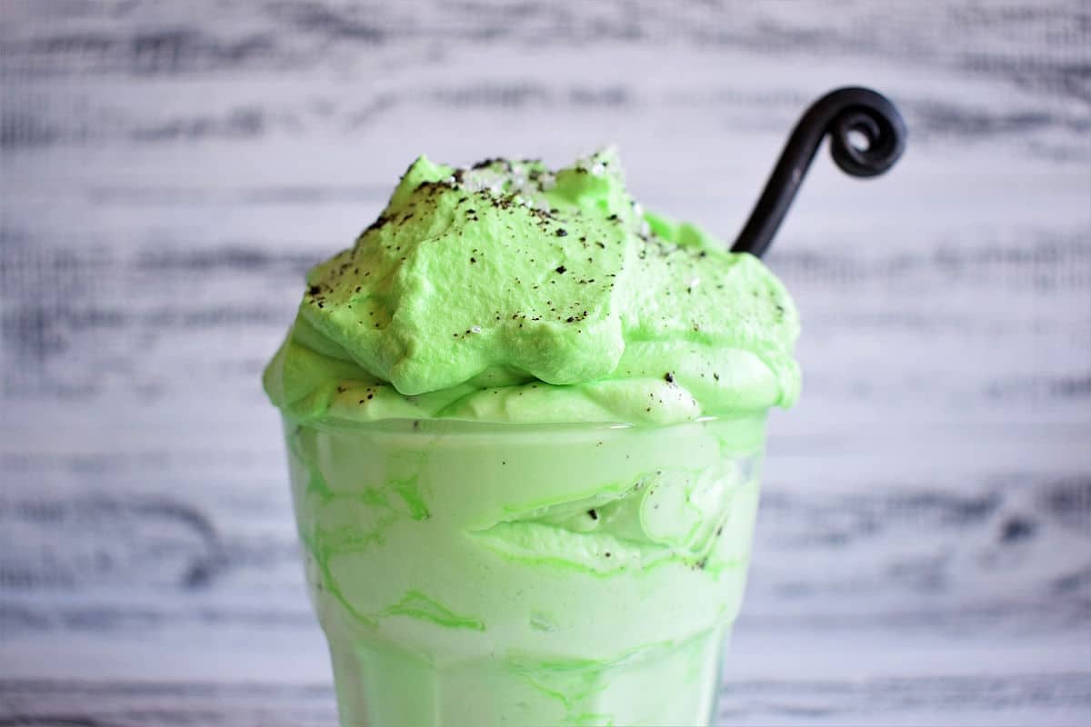 A glass of light green milkshake topped with fluffy green whipped cream, sprinkled with dark powder, and a black straw against a white wooden background.