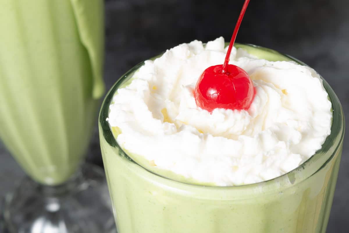 A green milkshake topped with whipped cream and a maraschino cherry, served in a clear glass.