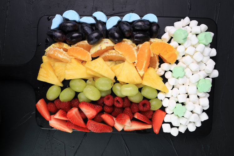 A rectangular tray displays rows of fruit and sweets: black grapes, orange slices, pineapple, green grapes, strawberries, raspberries, blue candies, and white mini marshmallows.