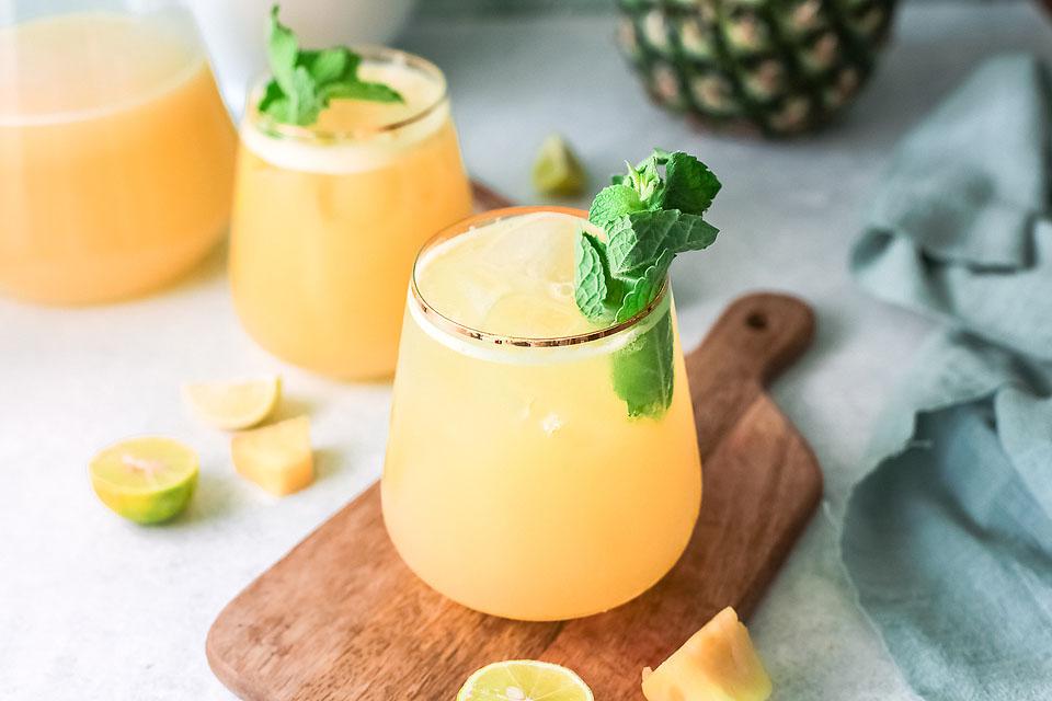 A glass of yellow tropical drink garnished with mint and a lime slice sits on a wooden board with lime wedges and pineapple pieces nearby. Another drink and a pineapple are in the background.