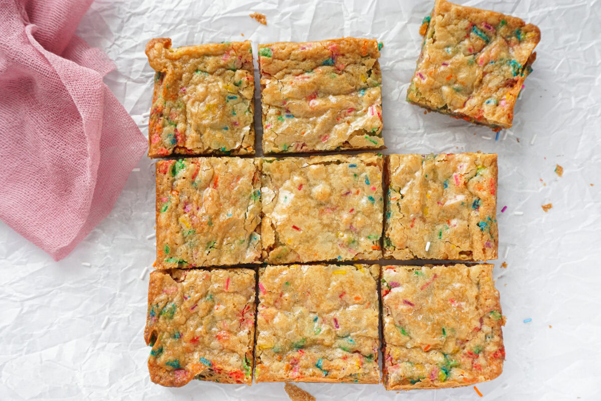 Nine funfetti blondie bars, one separated from the rest, are arranged on crumpled white parchment paper next to a folded pink cloth.