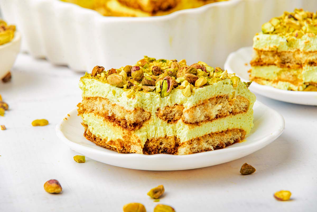 A slice of pistachio tiramisu with visible cream and ladyfinger layers, topped with chopped pistachios, on a white plate.