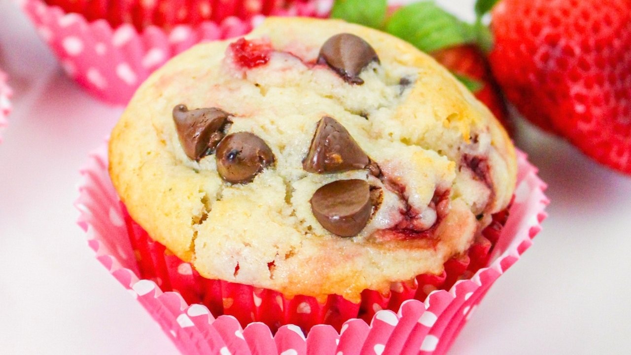 A strawberry muffin with chocolate chips in a pink polka dot paper wrapper, with fresh strawberries in the background.