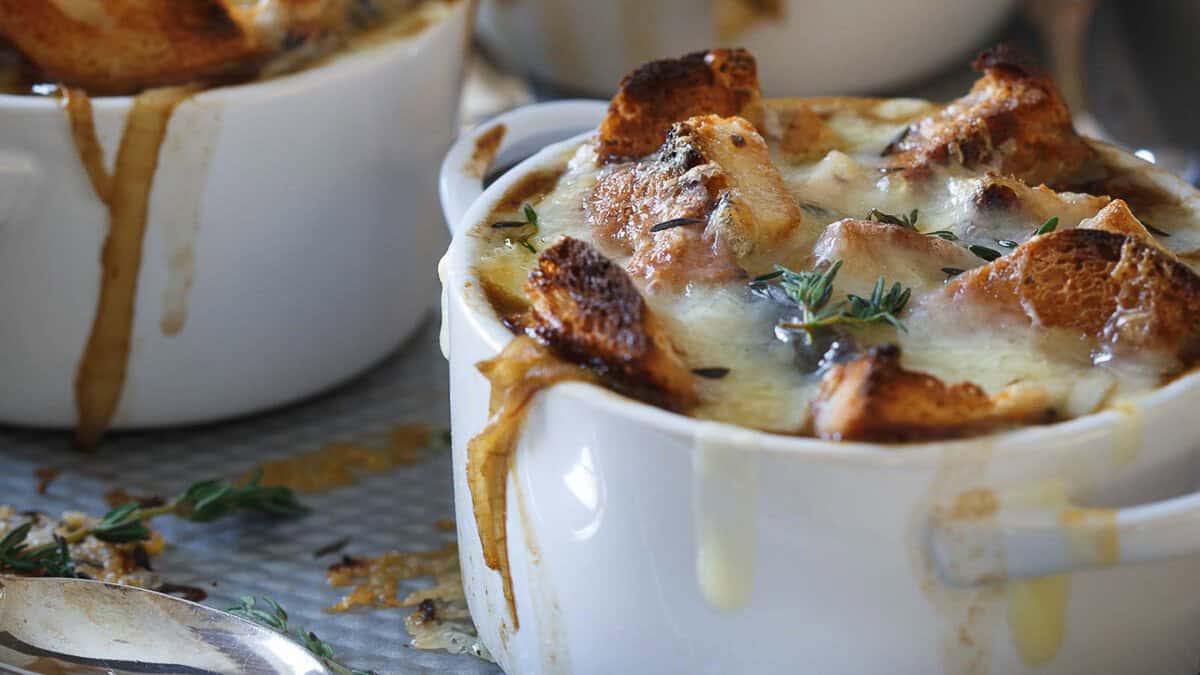 White ramekins filled with French onion soup, topped with toasted bread, melted cheese, and garnished with fresh thyme, with soup dripping down the sides.