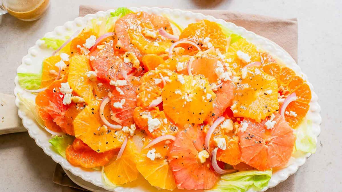 A platter of sliced oranges and grapefruits topped with crumbled cheese, thinly sliced red onions, black pepper, and lettuce on a white plate.