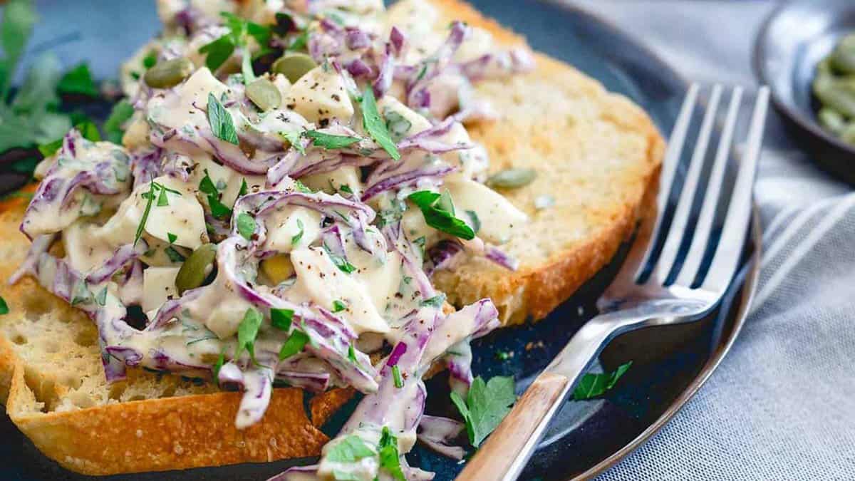 Two slices of toasted bread topped with creamy coleslaw, garnished with fresh herbs and seeds, served on a plate with a fork.