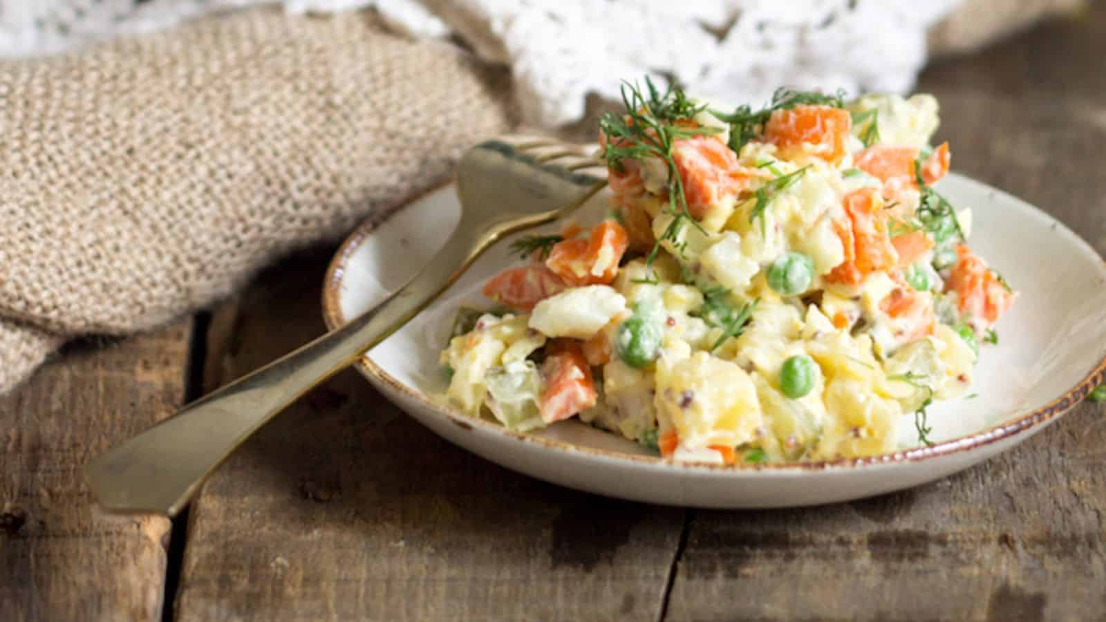 A plate of vegetable salad with carrots, peas, potatoes, and herbs, served with a gold fork on a rustic wooden table.