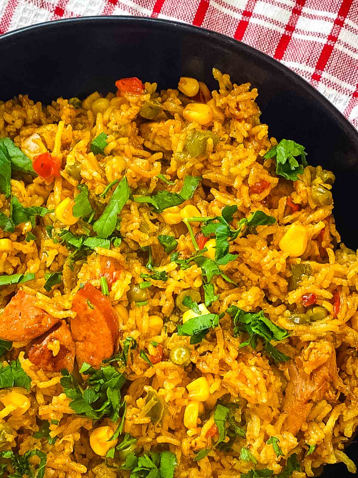 A close-up of a rice dish with sausage, corn, peas, carrots, and fresh chopped parsley in a black bowl on a red and white checkered cloth.