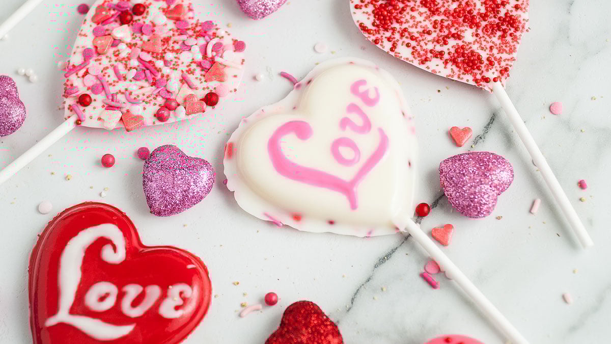 Heart-shaped lollipops decorated with the word Love and sprinkles, surrounded by glittery and plain candy hearts on a white surface.