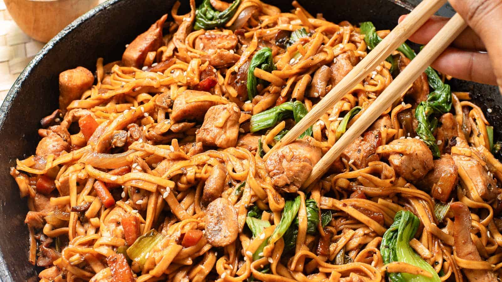 A skillet filled with stir-fried noodles, chicken, bok choy, and vegetables, with chopsticks picking up a portion.