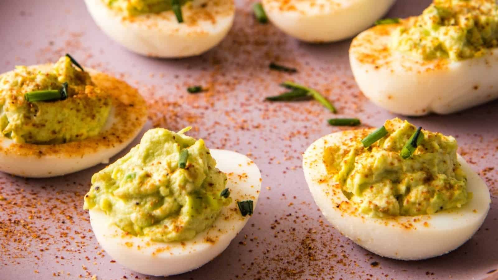 Halved hard-boiled eggs filled with a green, creamy mixture, garnished with chopped chives and sprinkled with red seasoning on a light surface.