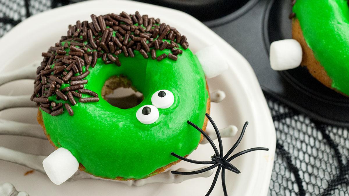 A donut with green icing, chocolate sprinkles, candy eyes, marshmallows for legs, and a plastic spider decoration on a white plate.