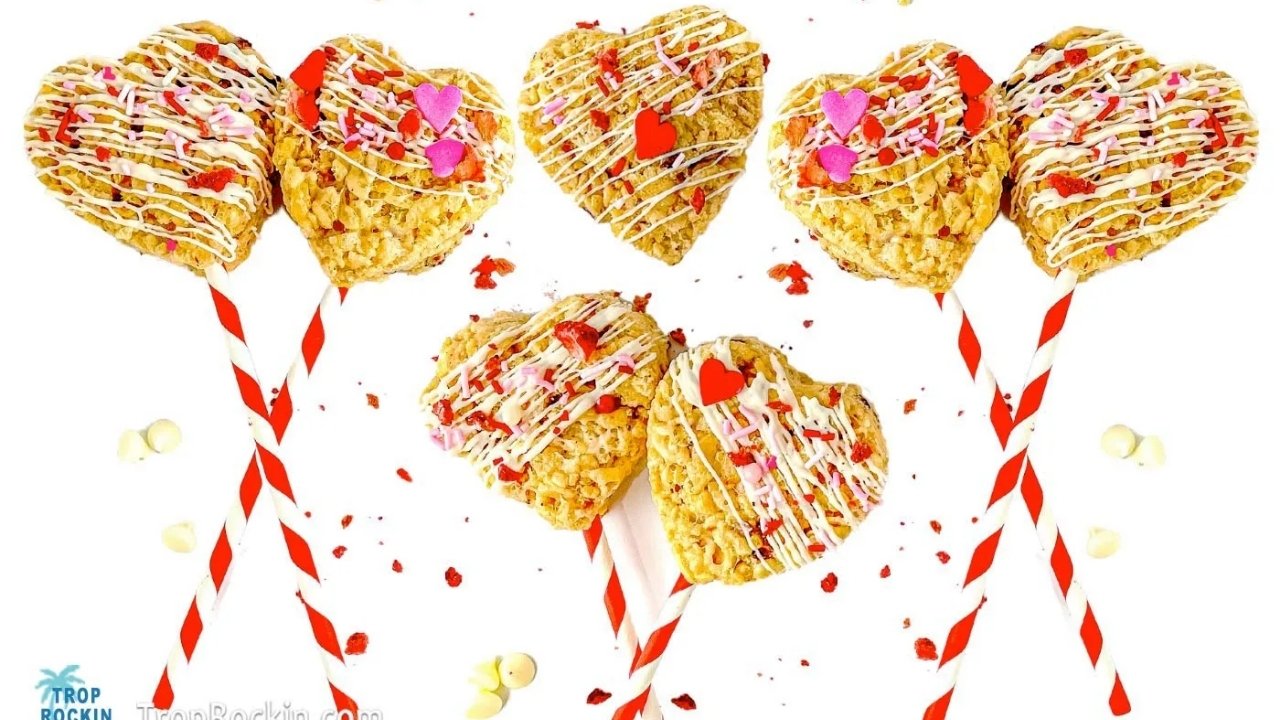 Five heart-shaped rice crispy treats on red-striped sticks, drizzled with white chocolate and decorated with colorful sprinkles and candy hearts, arranged on a white background.