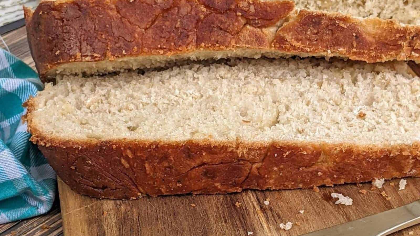 A close-up of a sliced loaf of bread with a golden-brown crust, resting on a wooden cutting board next to a blue and white cloth.