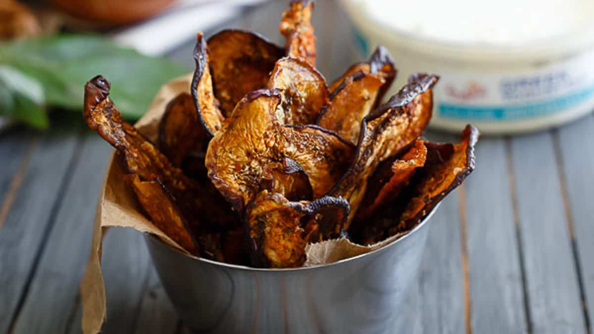A metal cup filled with crispy, browned eggplant chips lined with parchment paper on a wooden surface.