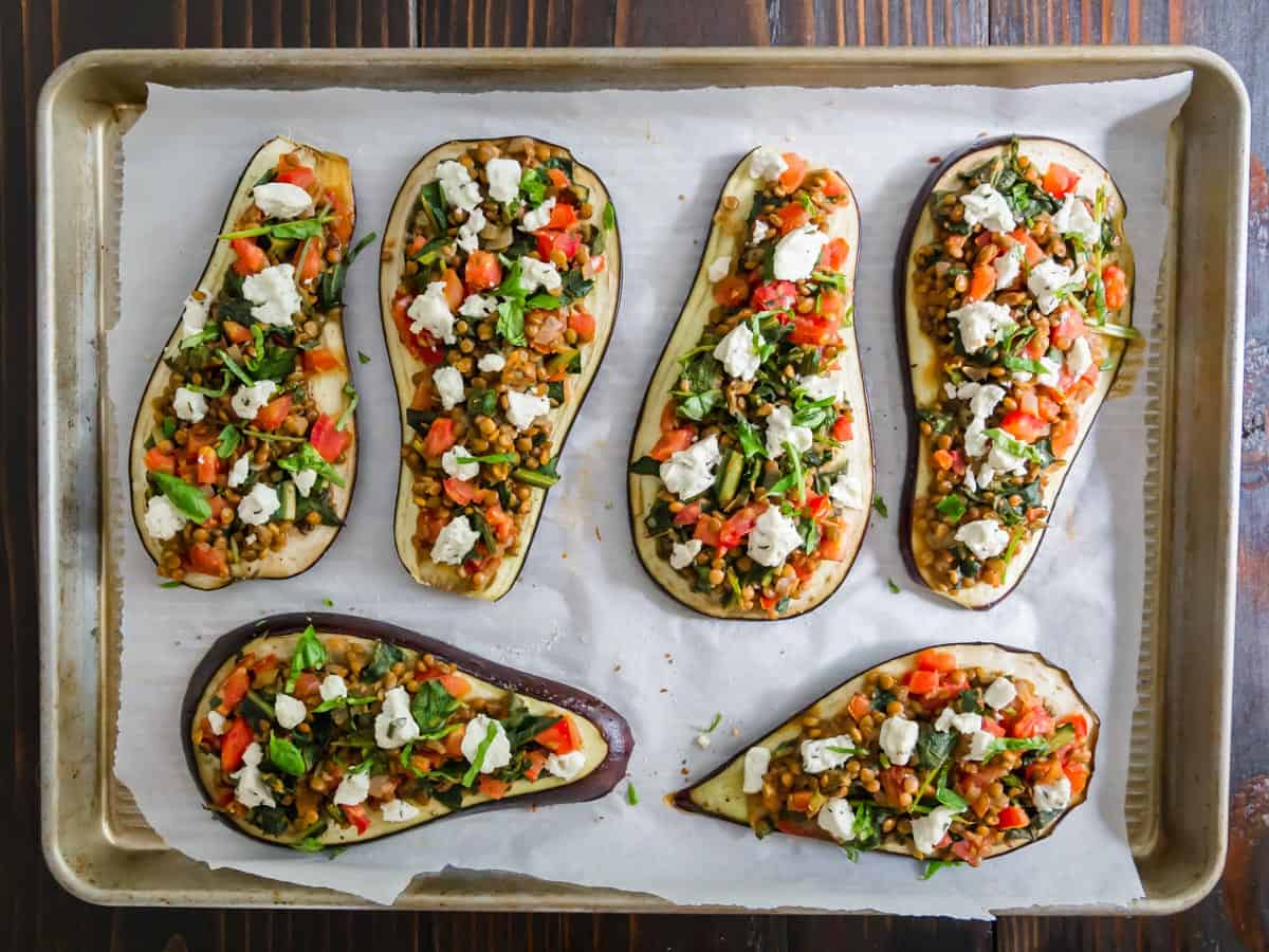 Six halved eggplants on a baking sheet, topped with a mixture of tomatoes, herbs, lentils, and white cheese, prepared for roasting.