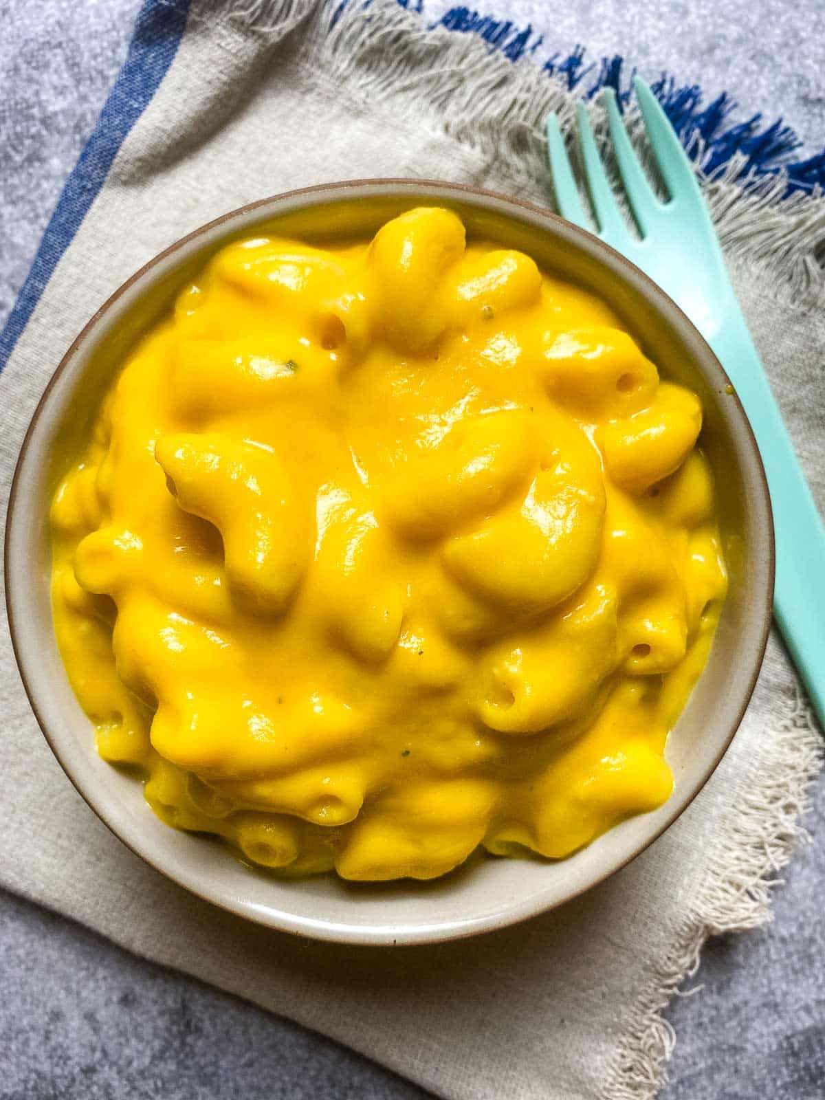 A bowl of macaroni covered in creamy, bright yellow cheese sauce, placed on a folded napkin with a light blue fork beside it.