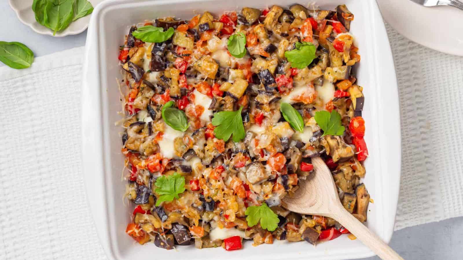 A white baking dish filled with baked ratatouille, containing diced eggplant, tomatoes, and peppers, garnished with fresh basil, with a wooden spoon resting inside.