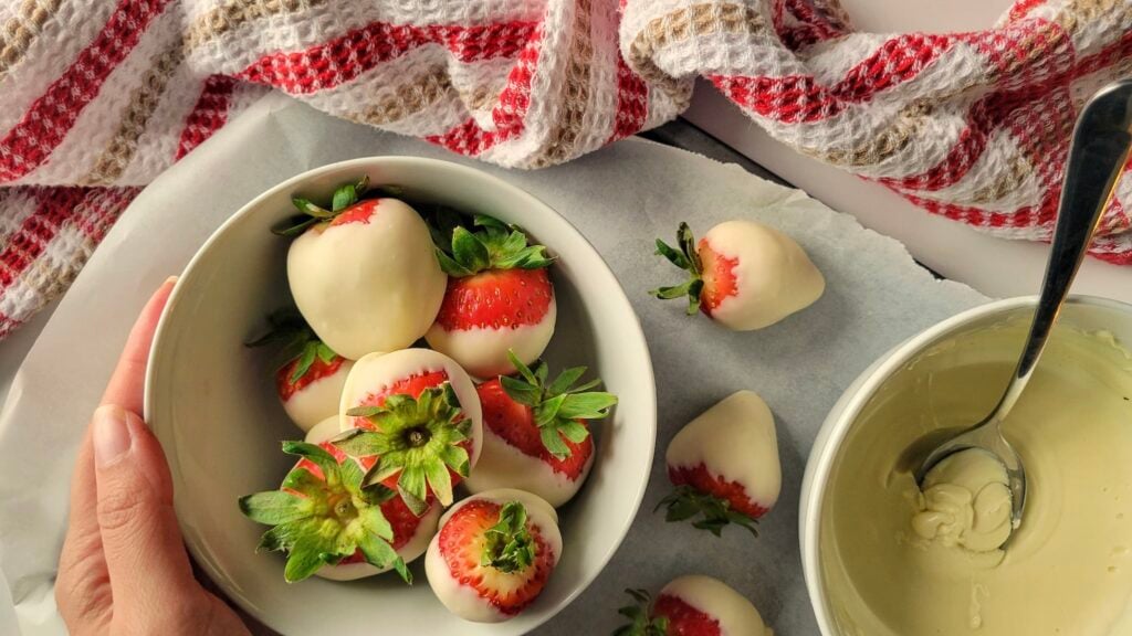 A hand holds a bowl of fresh strawberries dipped in white chocolate, with more dipped strawberries and a bowl of melted chocolate nearby on a table with a striped cloth.