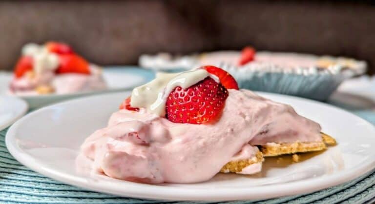 A slice of strawberry cream pie with graham cracker crust, topped with strawberries and whipped cream, served on a white plate.