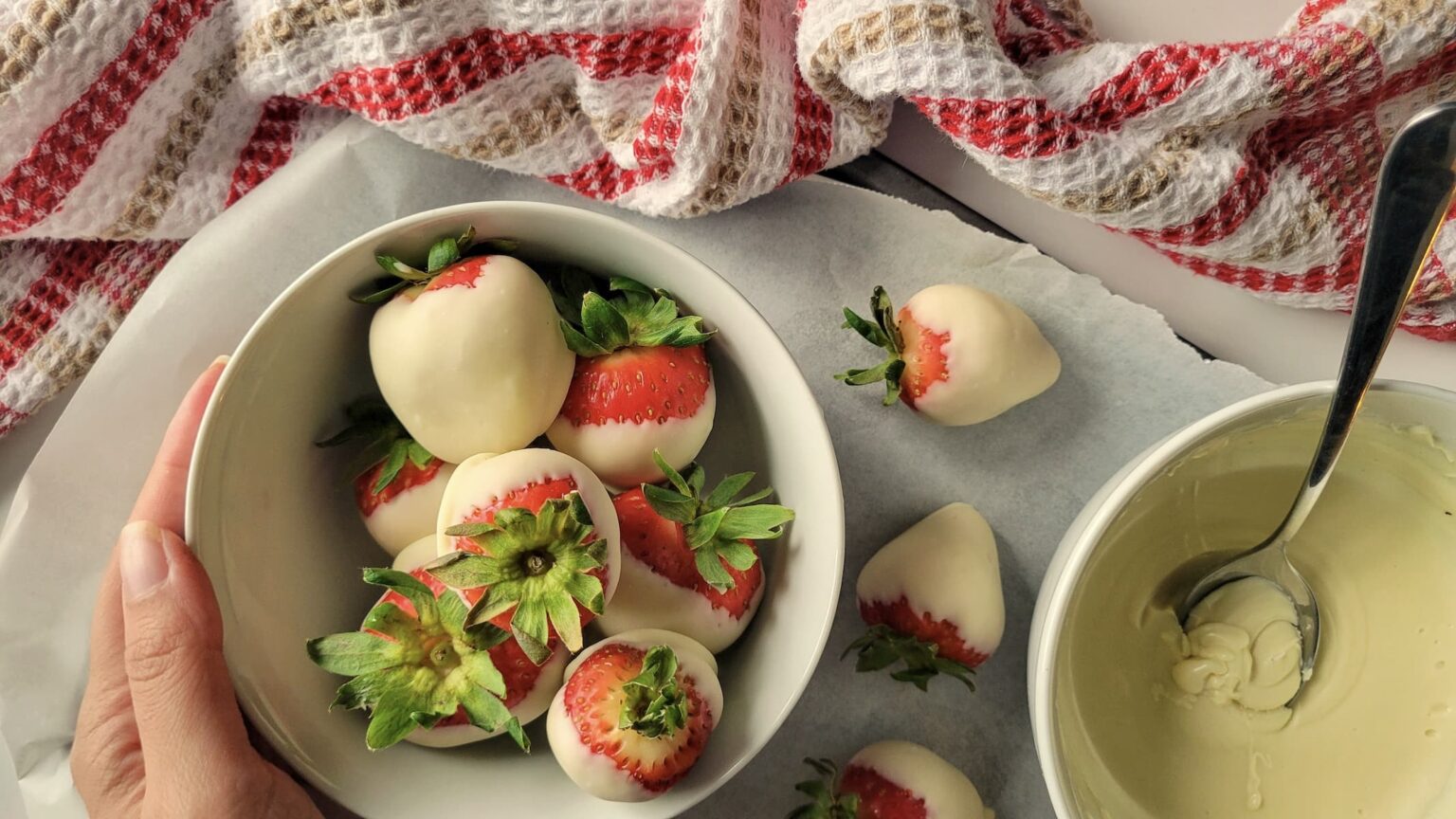 A hand holds a bowl of fresh strawberries dipped in white chocolate, with more strawberries and a bowl of melted white chocolate nearby on a parchment-lined surface.