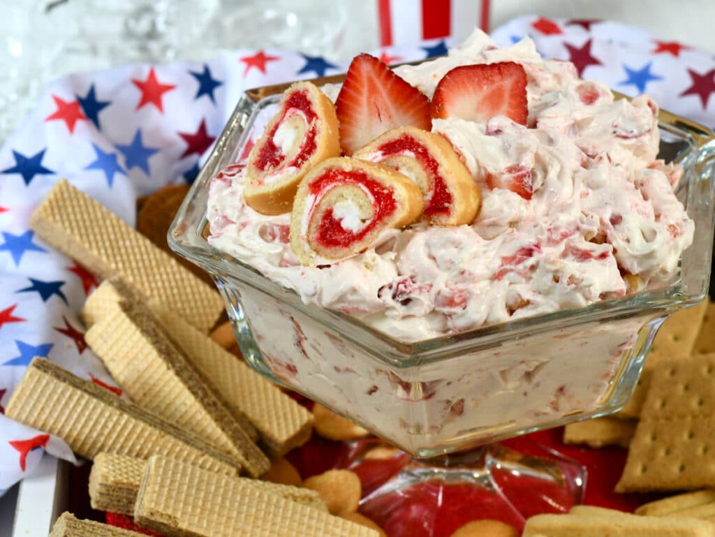 A glass bowl of strawberry cream dip topped with strawberry roll slices, surrounded by wafers, graham crackers, and cookies.