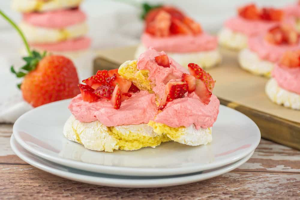A dessert with a meringue base, topped with pink whipped cream and chopped strawberries, is served on a white plate.