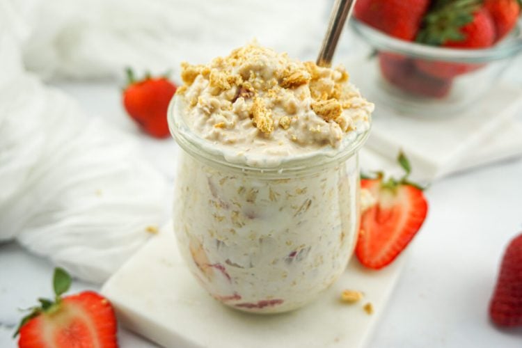 A glass jar filled with oatmeal mixed with strawberries and topped with granola, placed on a marble coaster, with whole and sliced strawberries nearby.