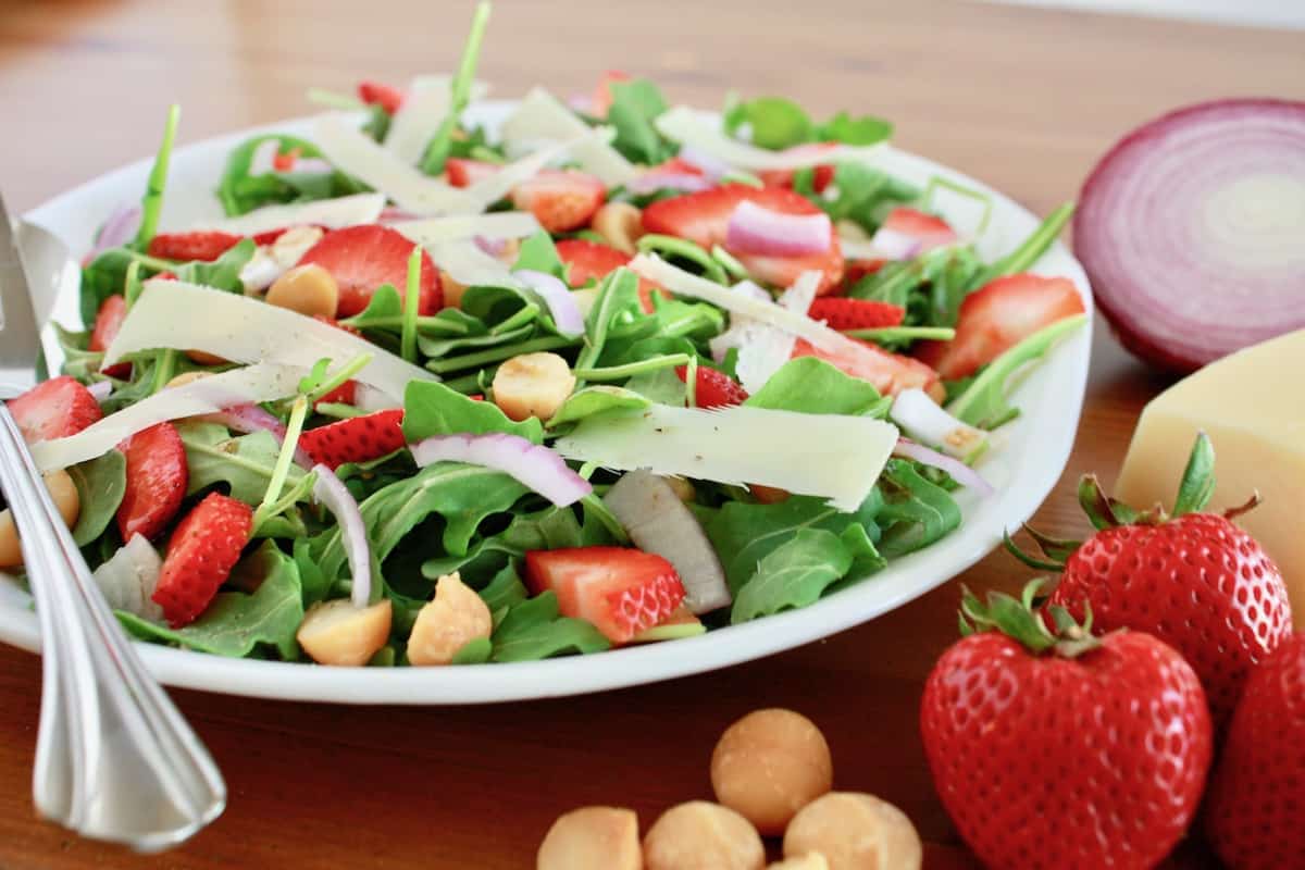 A plate of salad with arugula, sliced strawberries, red onion, macadamia nuts, and cheese shavings, with fresh strawberries and cheese on the side.