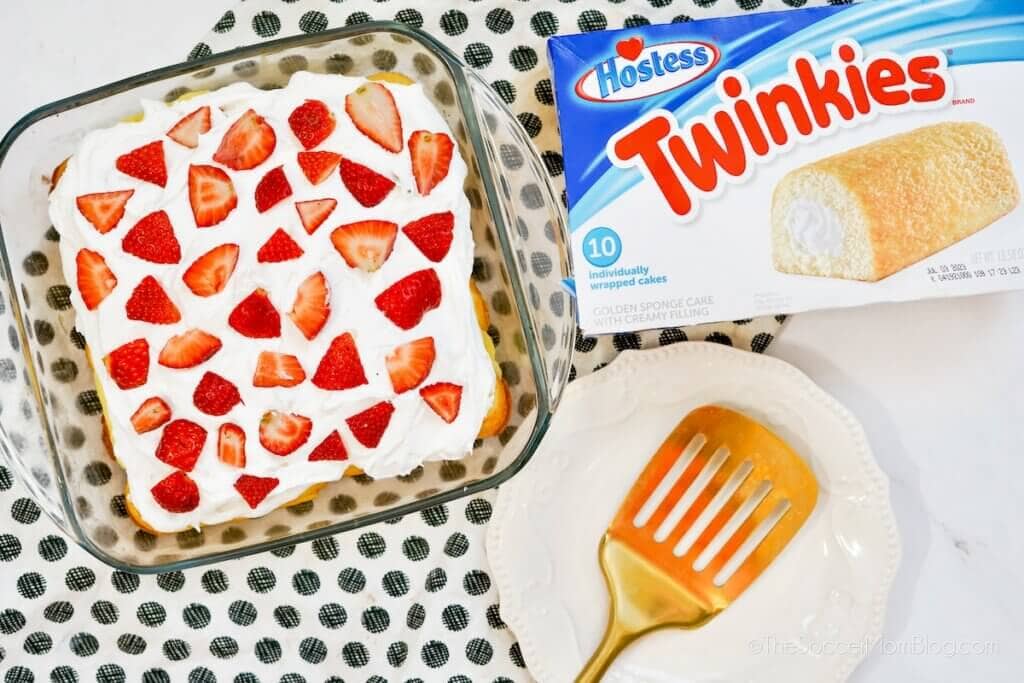 A glass dish of dessert topped with whipped cream and sliced strawberries sits next to a box of Twinkies, a gold spatula, and an empty plate on a patterned cloth.