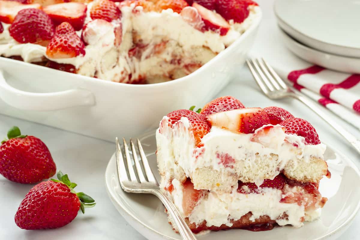 A serving of strawberry shortcake dessert topped with whipped cream and fresh strawberries on a plate, with a larger dish and whole strawberries in the background.