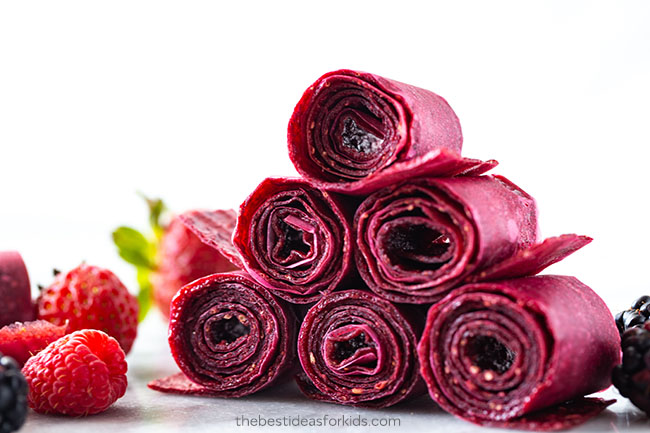 A stack of rolled homemade berry fruit leather is surrounded by fresh strawberries, raspberries, and blackberries on a white surface.