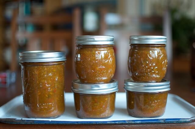 Six glass jars filled with orange preserve and sealed with metal lids are arranged on a white tray on a table.