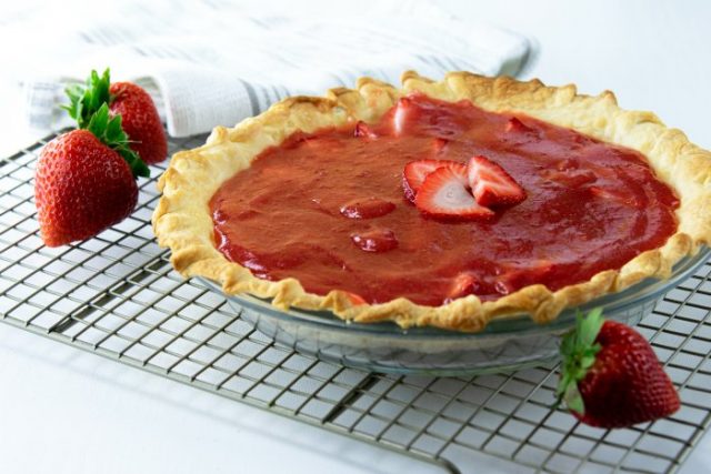 A strawberry pie with a golden crust and strawberry filling sits on a cooling rack, with fresh strawberries nearby and a striped towel in the background.