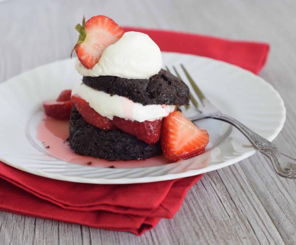 A chocolate shortcake dessert with sliced strawberries and whipped cream is served on a white plate with a fork, placed on a red napkin.