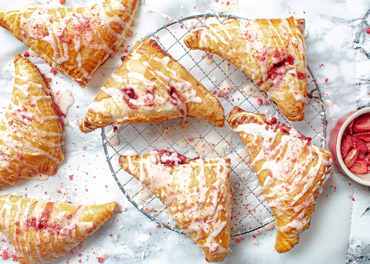Six triangular pastries with icing and strawberry filling are arranged on a cooling rack and marble surface. A small bowl with strawberry pieces is placed nearby.
